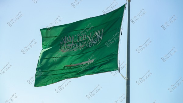 Image of the Saudi Arabian flag waving in the blue sky. The flag is green with white Arabic script of the Shahada in the center, beneath which is a white horizontal sword.