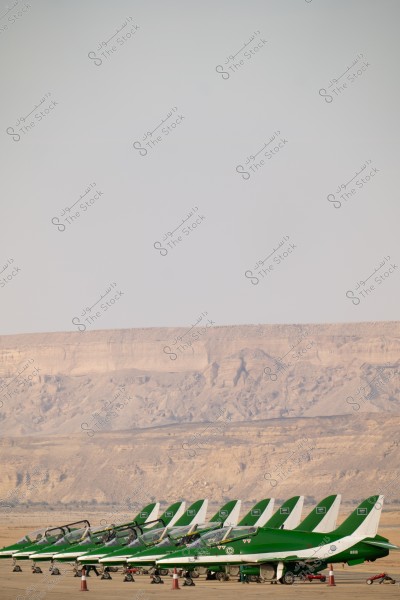 A lineup of green jet aircraft on a runway in a desert landscape. The planes feature Saudi national markings and are on a flat ground with sandy mountains in the background. The sky is slightly cloudy with natural lighting.