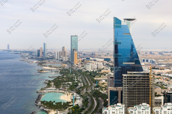 An image of Jeddah, Saudi Arabia, showcasing the beautiful waterfront. The photo features a modern blue skyscraper overlooking the ocean, with several other tall buildings along the coast. Sandy beaches and palm trees are prominently visible in the foreground, while the city stretches into the background.
