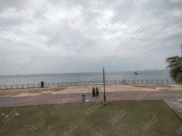 The image depicts a coastal walkway adjacent to the sea, with a few people walking along the pavement. The sky is overcast with clouds, and there is a lamp post and some greenery in the foreground. People are dressed in traditional clothing, suggesting the location might be in a Gulf region due to the attire and environmental style.