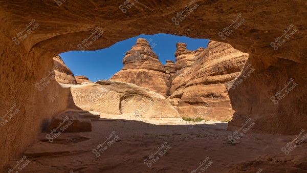 A natural landscape of massive sandstone rock formations in a desert creating an arch-like passage. The colors contrast between the reddish-brown of the sand and rocks and the clear blue sky. The details of the rock formations and their natural expressions are evident in the bright light.