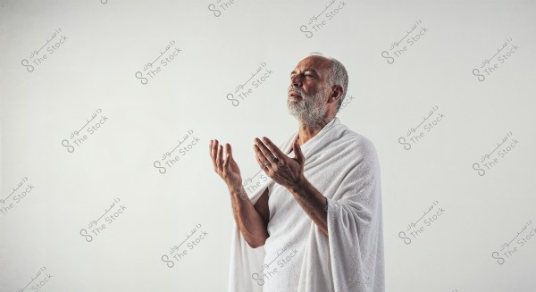 An image of an elderly man wearing a white Ihram, shown in a praying position with his hands raised. The background is white, highlighting the traditional Ihram clothing which may indicate Islam or the rituals of Hajj and Umrah.