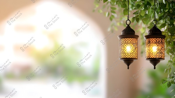 Two hanging lanterns with Arabic patterns illuminated by yellow light, surrounded by green foliage on a soft, blurred background.