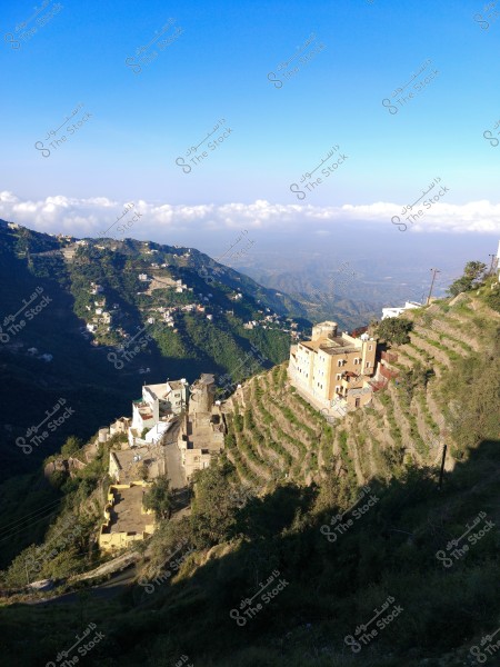 A view of a mountain village situated on a hillside, featuring white houses and terraced agricultural fields, beneath a bright blue sky.