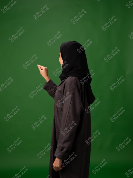 امرأة ترتدي حجابًا وعباءة سوداء تقف جانبًا على خلفية خضراء.