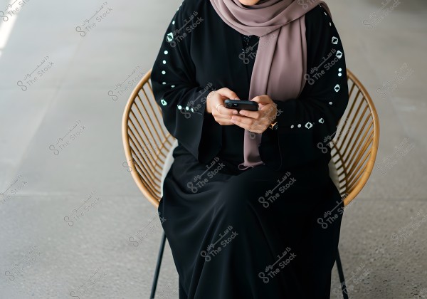 A woman sitting on a rattan chair, wearing a black abaya with simple designs on the sleeves, and a light brown hijab. She is holding a mobile phone in her hands, and the floor is made of gray tiles.