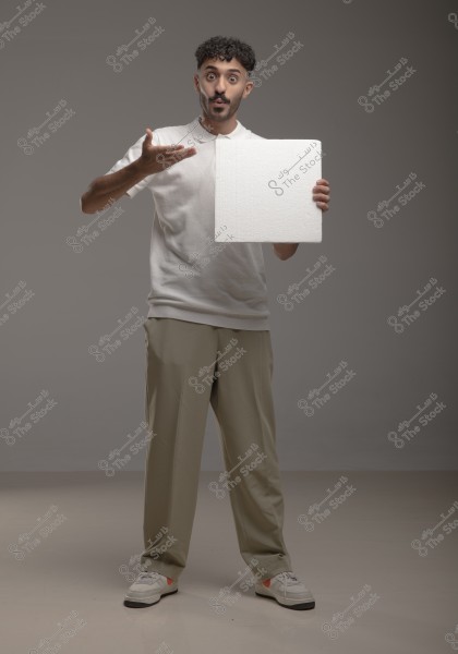 The image shows a young man standing in a studio wearing a white polo shirt, beige trousers, and sneakers. He holds a white rectangular piece in his right hand and gestures with his left hand. The background is a plain grey.