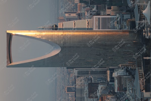 An image of the Kingdom Tower in Riyadh, Saudi Arabia. The tower rises high in the city skyline, featuring its metallic structure and distinctive inverted arch top. The tower reflects sunlight, surrounded by many other modern buildings, creating an advanced urban landscape.