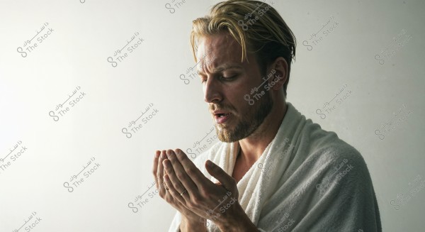 A portrait of a man with blonde hair and a light beard, wearing a white robe. He appears to be in a contemplative pose, with his hands together as if in prayer or meditation. The background is simple and uncluttered.