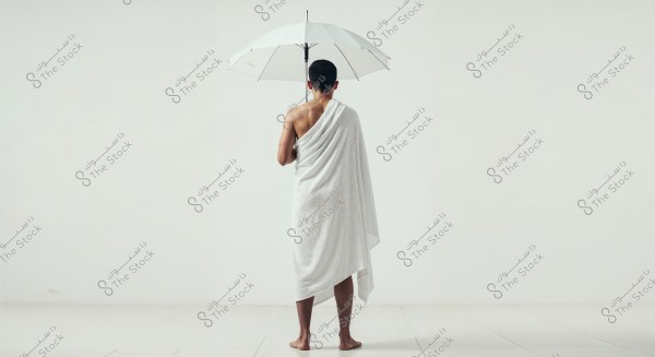 A photo of a man standing in a back view, wearing a white cloth draped around his body similar to Ihram. He holds a white umbrella above his head. The background is plain white, drawing attention to the person and his attire. The man is barefoot, standing on a glossy white surface.
