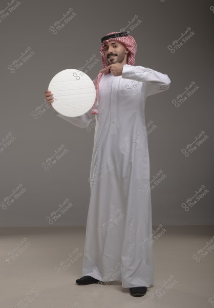صورة لرجل يقف مرتديًا الزي الخليجي التقليدي المكون من ثوب أبيض وغترة حمراء وعقال أسود. الرجل يمسك بيده قرصًا دائريًا أبيض ويشير إليه بيده الأخرى في وضعية مبتسمة أمام خلفية رمادية.