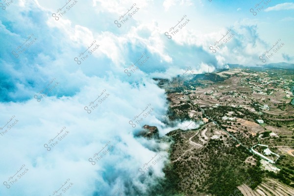 An aerial view of a stunning natural landscape featuring thick clouds covering mountains and green hills. The scene shows terraced agricultural fields in a spiral layout with scattered buildings amid green areas. The sky is blue with heavy clouds, adding an exceptional beauty to the scene.