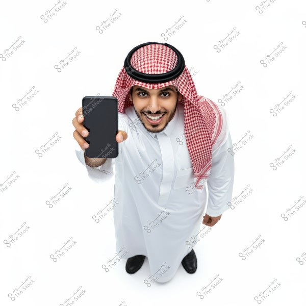 Image of a man wearing traditional Saudi attire, which includes a white thobe and a red and white checkered shemagh. He is smiling and holding a smartphone in front of him. The photo is taken from a top view angle.