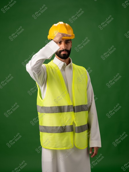 رجل عربي يرتدي خوذة أمان وسترة صفراء, يضع يده فوق عينيه للوقاية من الضوء, وخلفيته خضراء.