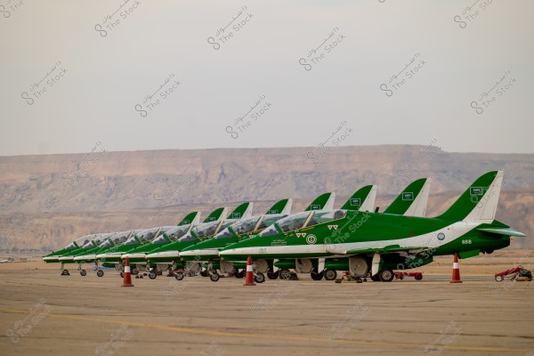 A lineup of green jet aircraft parked on a tarmac at a desert airport. The jets display Saudi Arabian markings and the Saudi flag on their tails and wings. A sandy mountainous backdrop is visible on the horizon beneath a clear sky.