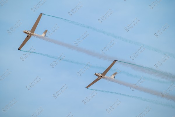The image shows two light aircraft flying in a clear blue sky, leaving colored smoke trails behind them. The aircraft are flying parallel to each other, emitting green and white smoke during an aerial display.