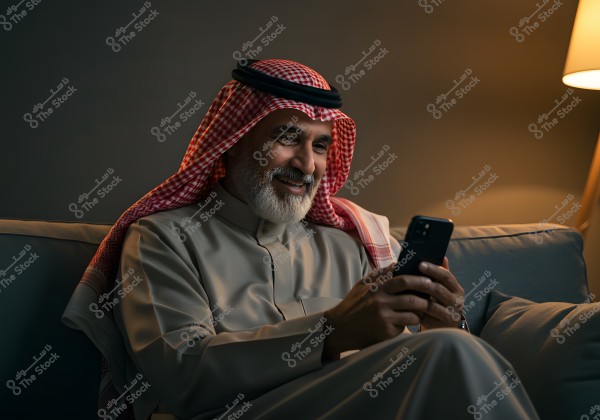 The image shows a man sitting on a couch, holding a mobile phone, and smiling as he looks at the screen. He is wearing traditional attire, including a thobe, a red and white checkered headscarf (shemagh), and an agal, suggesting he might be from a country like Saudi Arabia. The lighting in the room is warm, creating a cozy atmosphere.