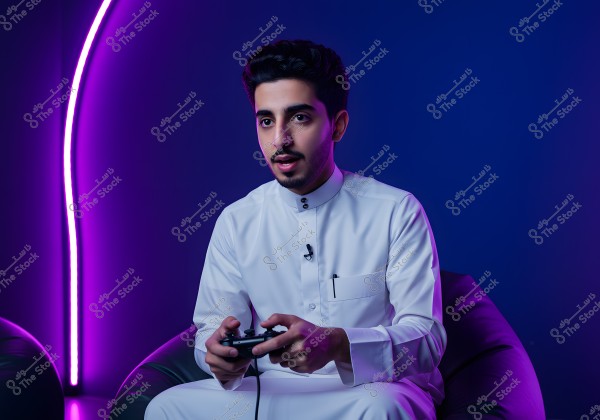 Image of a man sitting in a room lit with neon purple and blue lights, wearing a traditional white thobe that appears to be Gulf-style. He is holding a video game controller in his hands. The background features a curved neon light, providing a modern and contemporary feel.
