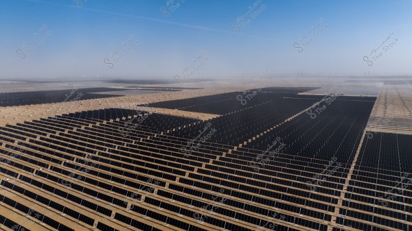 Image showing a vast field of solar panels arranged in neat rows on a flat desert landscape stretching as far as the eye can see. The field extends under a wide blue sky with a slight haze visible in the distant horizon.