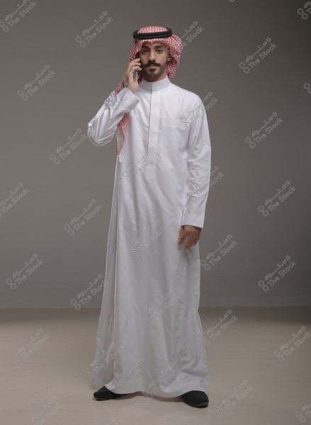 Image of a man standing in a light gray studio, wearing a white thobe and a red checked ghutra with a black agal, speaking on a mobile phone. The image conveys a sense of Gulf culture, with the clothing indicating Saudi origin.
