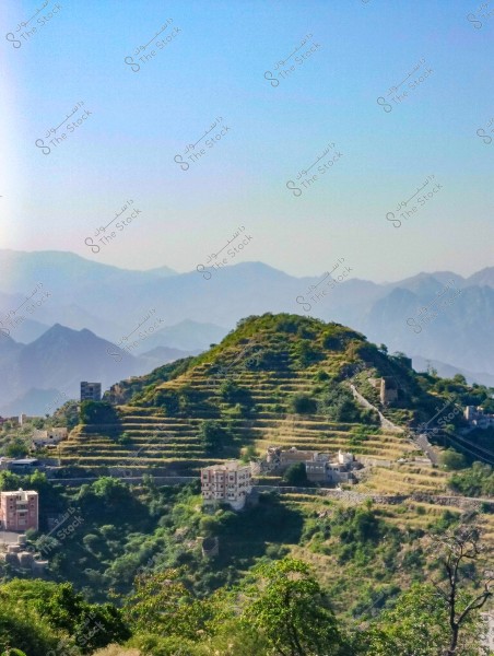 A scenic view of mountains with green agricultural terraces stretching across the hills. Several small buildings are scattered among the terraces. The mountains in the background appear hazy under the bright sunlight, with a clear blue sky above.