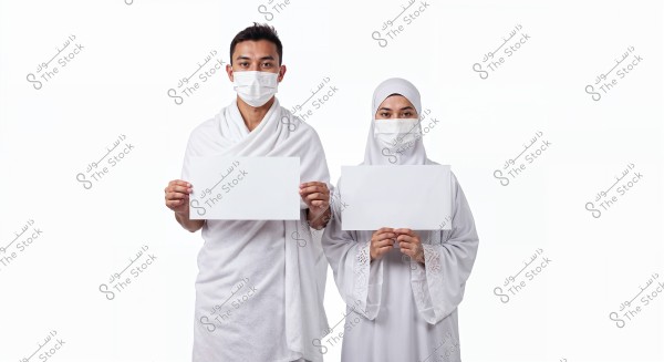 A man and a woman wearing white clothing and medical masks, holding blank white papers. The man is wearing an Ihram, and the woman is wearing a hijab and abaya.