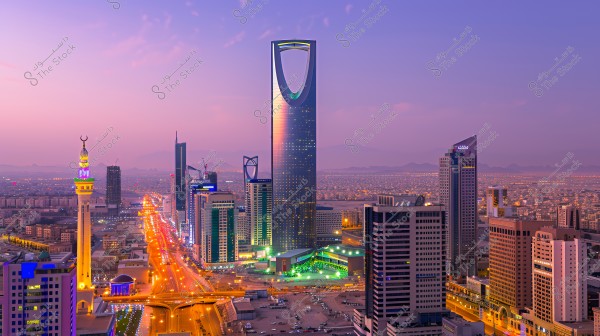 View of Riyadh city in Saudi Arabia at sunset. The iconic Kingdom Centre with its distinctive tower is prominent in the center, surrounded by tall buildings and illuminated streets. The horizon is a pink hue with mountains in the background.