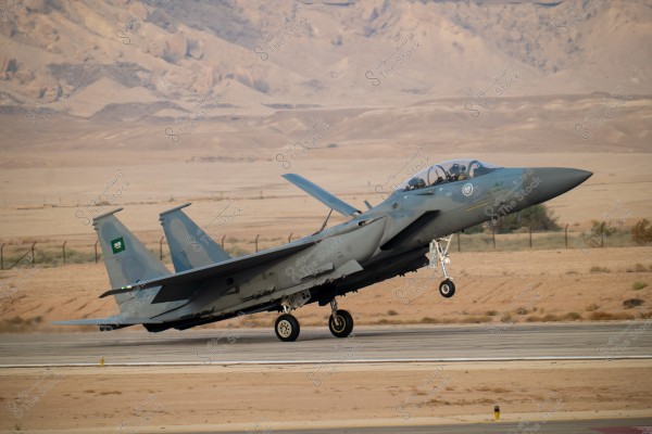 An F-15 fighter jet of the Royal Saudi Air Force taking off from a runway in a desert area. Desert sands and mountains are visible in the background. The Saudi flag is visible on the tail of the aircraft.