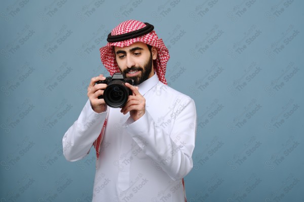 بورتريه شاب سعودي يرتدي الزي السعودي يحمل الكاميرا ويقوم بالتقاط الصور ، ويقف على خلفية سماوية