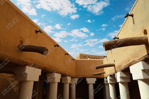 A view of a part of a traditional building under a blue sky with scattered clouds. The walls are made of natural clay with wooden beams extending from them, and white columns support the structure. The design reflects traditional Middle Eastern architecture.