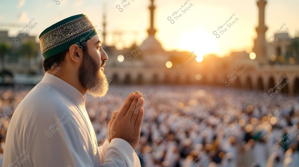 Side profile of a man wearing a white thobe and an embroidered cap with Islamic designs, raising his hands in prayer. Behind him is a crowd of people dressed in white in a large mosque courtyard, with minarets and traditional buildings in the background and a sunset on the horizon.