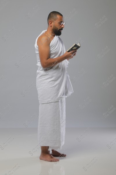 An image of a man standing in traditional white Ihram clothing, consisting of two pieces of fabric, holding a small open book. The man has a beard and is wearing glasses. The background is neutral with a light gray color, highlighting the details of the clothing and the person.