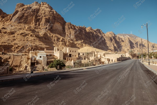 The image shows a newly paved road extending alongside a series of brown rocky mountains in the background. On the left side of the image, there are a cluster of traditional buildings made of stone and clay, accompanied by some green trees and vegetation. The contrast between the light brown color of the rocks and the clear blue sky above is prominent.