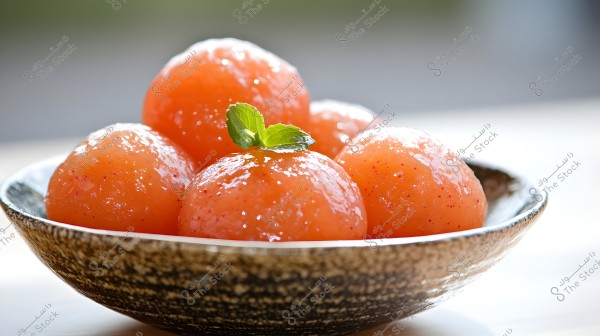 An image of a bowl containing shiny orange Gulab Jamun dessert balls, placed in a brown-designed bowl. The balls are topped with a mint leaf and appear appealing and vibrant. The background is blurred, making the bowl the focal point of the image.