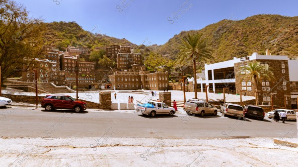 View of the historic Rijal Almaa village in Saudi Arabia, featuring traditional stone buildings with colorful windows. In the foreground, a paved road with parked cars and some people dressed in local attire standing beside the vehicles. Green hills surround the village under a clear blue sky.