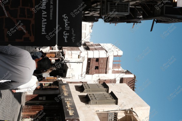 View of a street in Al-Alawi Market in Jeddah. The historic buildings with traditional facades and shop signs are visible, with several people walking in the street.