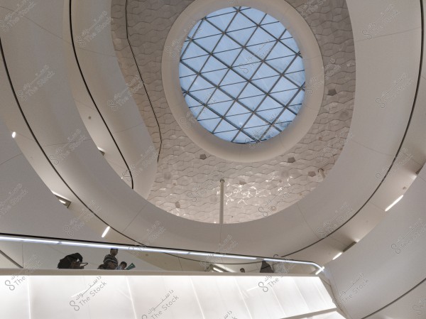 The image shows a modern interior design of a building featuring a large circular glass ceiling with a geometric grid, allowing light to enter the surrounding space. White arches frame the opening, with geometric details on the walls. Several people are visible on the lower floor, looking upwards and seemingly interacting with the architectural space.