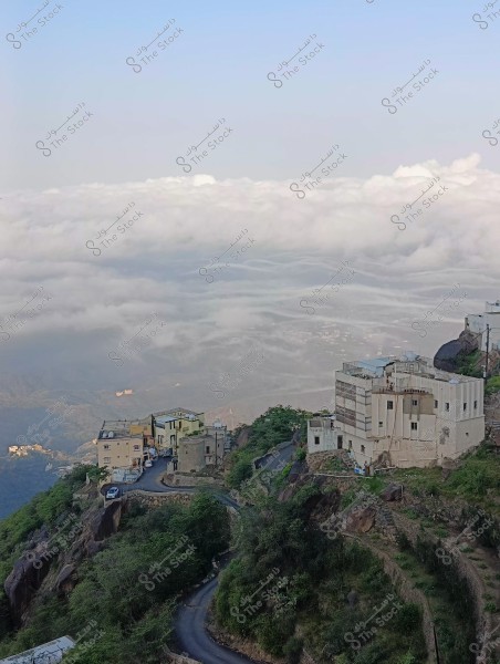 منظر طبيعي مدهش يطل على قرية على قمة الجبل, مع طريق متعرج يؤدي إلى مجموعة من البيوت التقليدية المحاطة بالخضرة. تحت السحب الكثيفة التي تغطي الوديان, يمكن رؤية البيوت مبنية بالحجارة والأسمنت, ويظهر في الخلفية سماء صافية.