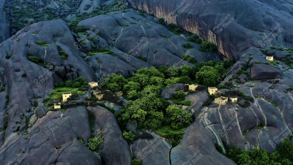 Aerial view of small traditional houses nestled among rocky cliffs in a mountainous area. The rocks appear dark, while green trees and vegetation fill the crevices and spaces between the rocks, forming a layered and beautiful natural landscape.