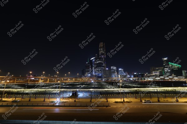 A nighttime cityscape featuring tall, illuminated skyscrapers with a wide road in the foreground and a lit street bridge. The buildings have modern architectural designs, with some displaying colorful lights that stand out at night.