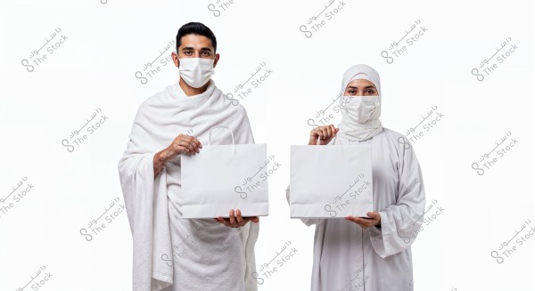 A photo of a man and woman wearing white Ihram garments, holding white shopping bags. They are wearing medical masks. The woman is wearing a hijab that covers her entire head. The background is white.