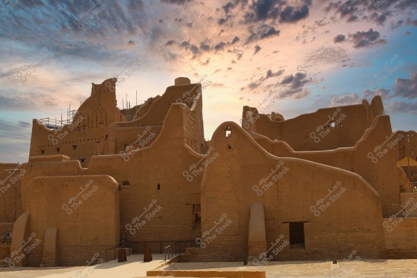 An image of ancient mudbrick ruins representing traditional architecture in Diriyah, Saudi Arabia. The brown mudbrick walls are set against a sky filled with clouds during sunset, casting a warm light over the scene.