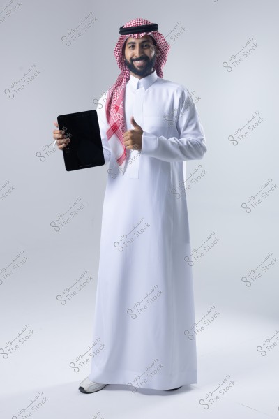 Image of a man wearing a white thobe with a red shemagh and an agal, standing against a white background. He is smiling and holding a tablet in one hand while giving a thumbs-up with the other hand. The clothing suggests a traditional Saudi style.