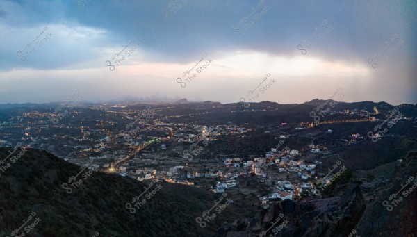 A panoramic view of a city lit at night stretching across a valley surrounded by mountains. The sky is cloudy with faint sunset light on the horizon. The illuminated lights display the distribution of buildings and streets in the city, with several lit roads extending through the mountains.