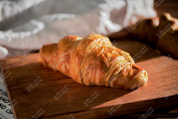 The image shows a golden croissant placed on a wooden board. The croissant features a thin, crispy crust. In the background, there\'s a partially visible white cloth, adding a warm and cozy feel to the image.