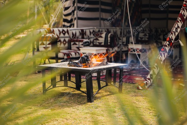 A scene of an outdoor seating area featuring a traditional tent with furniture adorned with Arabic motifs and patterned fabrics. In the foreground, there is a lit fire pit on a metal table, surrounded by a grassy ground and a backdrop with colorful tribal woven patterns.