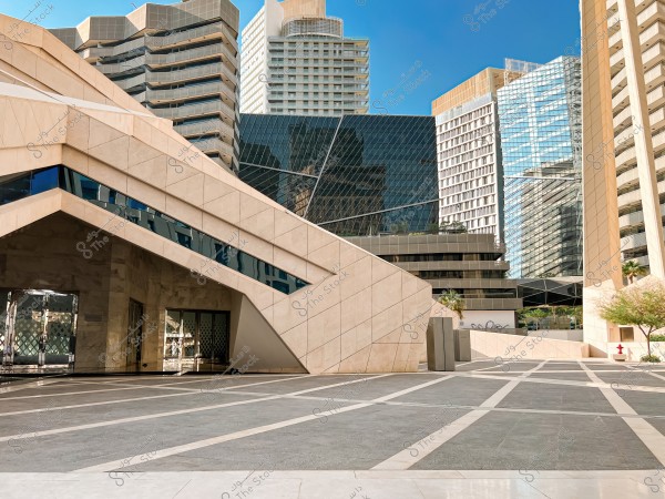 Image of a modern complex featuring multiple buildings with contemporary architectural style. The front building has a slanted design and is covered with tiles and glass panels, surrounded by other tall buildings with various shapes and designs, made of glass and tiles. A wide plaza with a geometric floor design is seen in the foreground. Trees and plants are visible in the surrounding areas.