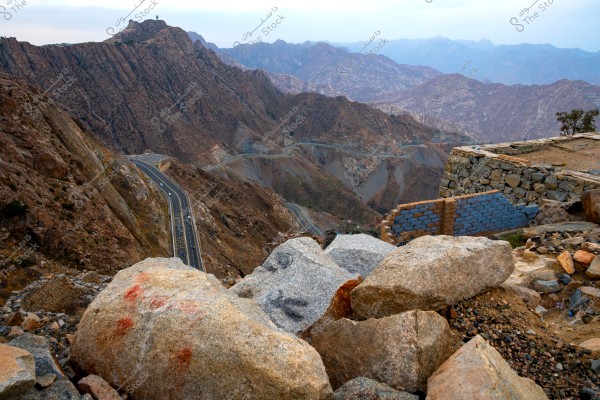 The image shows a natural landscape of rugged mountains with a modern asphalt road winding through them. Large rocks are scattered in the foreground, and some stone walls are built alongside the road. The background features a range of successive mountains under an open sky.