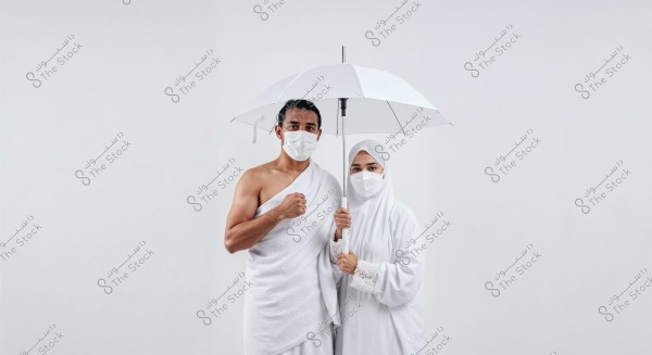 A portrait image of a man and woman on a white background. The man is wearing a white Ihram covering his left shoulder, and the woman is wearing a white abaya and hijab. Both are wearing white medical masks and holding a white umbrella above their heads. The image reflects the atmosphere of Hajj.