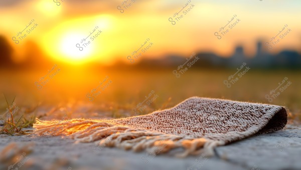 A small prayer rug placed on the ground during sunset. The rug has a textured fabric with fringes on the sides. The horizon in the background is blurred, with golden and orange hues of the setting sun filling the sky.